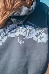 Hoodie gris con capucha y cordones ajustables, de corte holgado y manga larga. Presenta un bordado de flores blancas en la espalda y en el puño.