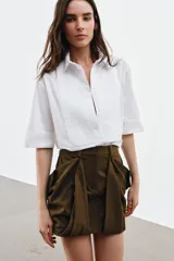 Camisa de algodón con cuello solapa y mangas por debajo del codo. Presenta un diseño con pechera plisada frontal, bajo recto y cierre mediante botones ocultos bajo solapa.