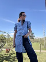 Camisa tipo vestido a rayas celestes, rojas y blancas, con cuello alto fruncido, mangas cortas abullonadas con puños elásticos y cierre frontal de botones.