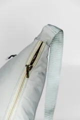 Bandolera color gris claro con forma de media luna, cierre de cremallera y correa ajustable. Incluye etiqueta metálica con el logo de la marca.