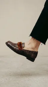 Mocasín de cuero con textura gastada, color marrón, con detalle de pelo en la parte superior y suela plana.