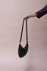 Bolso de hombro tipo hobo, confeccionado en gamuza color marrón oscuro, decorado con tachas metálicas plateadas en toda su superficie frontal. Cuenta con un asa corta fija y cierre superior.