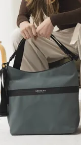 Cartera tipo hobo de tamaño grande en color beige, con detalles en negro. Presenta una franja horizontal negra con el logo de la marca, asa corta de hombro y correa larga desmontable. Incluye bolsillo frontal con cierre oculto y bolsillo trasero.
