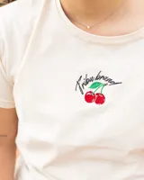 Remera de jersey de algodón color crudo, con cuello redondo y mangas cortas. Presenta bordado de cerezas y texto en el frente.