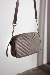 Cartera bandolera color gris topo, con diseño capitoneado en forma de V en el frente, correa de cuero con cadena metálica, cierre metálico y doble bolsillo interior.