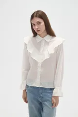 Camisa de algodón blanca con cuello camisero, cierre frontal de botones y volados bordados en el frente y hombros. Presenta mangas largas con puños acampanados y detalles bordados en el ruedo.