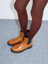 Bota tipo chelsea de cuero con textura de reptil, color suela. Presenta paneles elásticos laterales en color negro, suela de goma dentada con pespuntes en contraste y puntera redondeada.