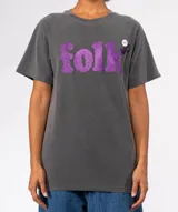 Remera de algodón color gris oscuro con lavado especial, corte holgado y cuello redondo. Estampa frontal con la palabra "folk" en glitter morado. Lleva un pin decorativo en la zona del hombro.