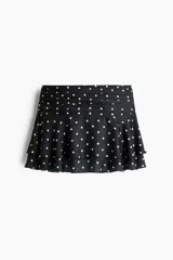 Pollera mini de satén negro con estampado de lunares blancos. Presenta una cintura ancha con pliegues horizontales y un diseño escalonado con volados que aportan volumen y movimiento.