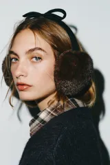 Orejeras tipo diadema con estructura de vincha negra y detalle de lazo en la parte superior. Presentan almohadillas con textura de pelo sintético en color marrón.