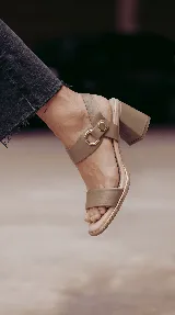 Sandalia de cuero color taupe con taco cuadrado de 6 cm, tira frontal ancha y tira en el tobillo con detalle metálico dorado y hebilla circular.