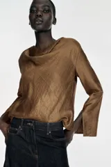 Blusa de manga larga con cuello fluido tipo drapeado y abertura en los puños. Presenta un acabado satinado con textura y cierre posterior con cremallera oculta.