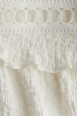 Blusa blanca de manga larga con cuello alto y lazo para atar. Presenta detalles de volados y calados tipo encaje en el canesú y puños con elástico y volado.