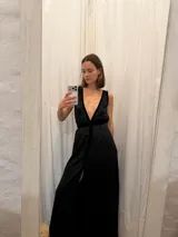 Vestido largo de seda color bronce con acabado brillante, escote profundo en V y espalda abierta. Presenta detalles en terciopelo negro que enmarcan el escote, recorren el frente y definen la cintura con una faja ajustable. Posee breteles anchos y una abertura frontal en la falda.