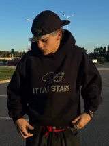 Hoodie de color negro con diseño de estampa frontal que combina una tipografía manuscrita con el texto ITTAI STARS en letras mayúsculas. Presenta un corte holgado, capucha amplia y bolsillo tipo canguro.