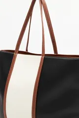 Cartera tipo tote bag en símil cuero, con diseño en bloques de color negro y blanco, y detalles en marrón. Tiene asas largas para llevar al hombro y un cierre magnético.