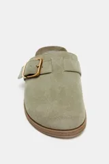 Zueco plano de serraje color verde oliva, con punta redonda y detalle de tira con hebilla metálica dorada en el empeine. Cuenta con plantilla de efecto aterciopelado y suela de corcho.