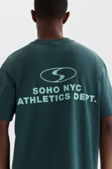 Remera boxy de punto liviano de algodón en color verde oscuro, de hombros caídos, con cuello redondo acanalado y bajo recto. Presenta un diseño estampado en el pecho con texto y logotipo.