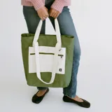 Bolso tote verde militar confeccionado en nylon resistente al agua, con un bolsillo frontal grande en color crudo que incluye cierre horizontal y una anilla metálica. Cuenta con asas largas de hombro en color crudo y una manija superior corta.