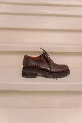 Mocasín de cuero vacuno negro con textura tipo reptil, suela track de goma, cordones finos y solapa con flecos sobre el empeine.