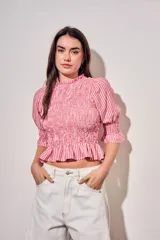 Blusa de algodón con diseño a rayas verticales en tonos rosa y blanco. Presenta un cuerpo con fruncido elástico (smock), cuello alto con volados y mangas tres cuartos abullonadas con terminación en volado.