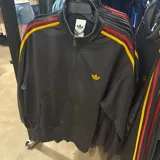 Campera deportiva Adidas con cierre frontal, cuello alto y bolsillos laterales. Presenta diseño en color negro con las icónicas tres tiras en los hombros en tonos rojo y amarillo, y logo del trébol bordado en amarillo en el pecho.