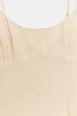 Vestido corto estilo lencero color beige, con escote redondo, tirantes finos ajustables y detalles de encaje y bordados a tono. Presenta un diseño con costura horizontal en la cintura y bajo acabado con volantes.