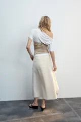 Falda midi de tiro medio color gris, con diseño asimétrico en el bajo, bolsillos frontales y traseros, y cierre frontal con cremallera y botón.