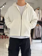 Campera tipo canguro con cierre frontal completa y capucha, de corte boxy fit en color blanco marfil. Presenta bolsillos frontales tipo parche y puños y dobladillo en rib elástico.
