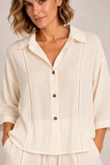 Camisa de lino color crudo, con cuello camisero, mangas tres cuartos y cierre frontal de botones marrones. Presenta detalles de costuras caladas verticales en el frente.