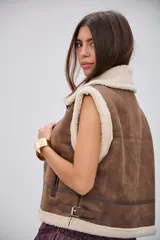 Chaleco de estilo aviador en color camel, confeccionado en material efecto cuero con interior y bordes de corderito sintético color crema. Presenta cierre frontal, cuello solapa y detalles de hebillas metálicas en los laterales y el cuello.