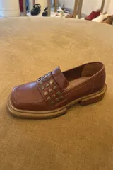 Mocasín de cuero color suela con punta cuadrada y detalle de tachas metálicas en la faja frontal. Cuenta con suela de caucho con vira a la vista.