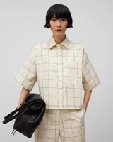 Camisa de algodón color crema con estampado de cuadros negros. Tiene cuello solapa, manga corta, bolsillo en el pecho y cierre frontal con botones ocultos.