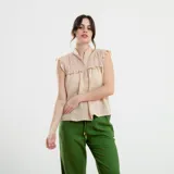 Camisa sin mangas color beige, con cuello alto con volados, detalles de encaje calado en los hombros y parte superior del pecho, y cierre frontal con botones.