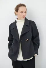 Chaqueta de paño color gris oscuro, con solapas amplias y cierre frontal mediante un alamar único. Presenta bolsillos laterales y mangas largas con puños rectos.