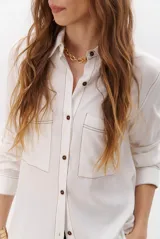 Camisa beige de manga larga, con cuello clásico, botones marrones y dos bolsillos frontales. Presenta costuras decorativas en contraste.