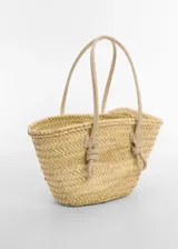 Bolso tipo shopper de fibra natural de hoja de palma, con diseño trenzado y detalles en piel bovina. Cuenta con dos asas largas con nudos decorativos y cierre de gancho.