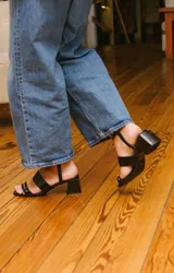 Sandalias negras de cuero con dos tiras horizontales sobre el pie y pulsera al tobillo con ojalillos y tachas. Taco cuadrado de 5 cm.