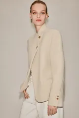 Blazer de corte estructurado en color beige, con cuello mao y cierre frontal cruzado mediante botones. Presenta mangas largas con detalle de botones en los puños y bolsillos de vivo en el frente.