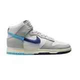 Championes Nike Dunk High Retro SE de corte alto, con diseño en paneles de cuero blanco y gris. Presenta un swoosh lateral dividido en dos tonos de azul, suela intermedia con acabado envejecido y suela exterior bicolor en azul claro y oscuro.