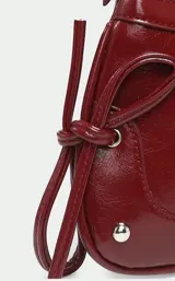 Cartera tipo baguette de charol color bordó, con diseño estructurado, cierre superior y detalles de tachas metálicas en las esquinas inferiores. Incluye tiras laterales decorativas y correa de hombro ajustable.