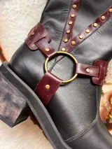 Bota alta de cuero negro con taco cuadrado de efecto madera, puntera redondeada y detalles de tiras en tono bordó con tachas metálicas doradas. Cuenta con una argolla metálica en el empeine y una hebilla de ajuste en la parte superior de la caña.