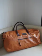 Bolso de viaje tipo weekender de cuero color tostado con asas y detalles en marrón oscuro, herrajes metálicos y bolsillo frontal con cierre.