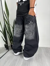 Pantalón de mezclilla negro de corte muy ancho (baggy), con efecto de lavado tipo 'acid wash' o 'dip-dye' que aclara la parte superior de los muslos. Presenta bordado decorativo en el bolsillo trasero derecho con forma de cruz rodeada de cuentas y tachas plateadas.