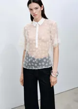 Camisa de manga corta con diseño recto y cuello camisero. Confeccionada en tejido semitransparente con bordado de flores pequeñas y cierre frontal con botones.