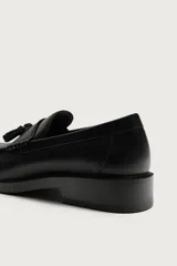 Mocasín de vestir color marrón oscuro, confeccionado en cuero, con detalle de antifaz y borlas decorativas en el empeine. Presenta una horma redonda y suela plana a tono.