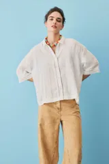 Camisa blanca de lino, con mangas anchas a tres cuartos, cuello clásico, cierre frontal con botones y detalles de calados verticales en la parte central y en los puños.