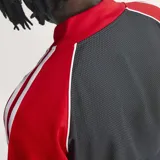 Campera deportiva Adidas SST de corte holgado, confeccionada en tejido de malla color negro con mangas rojas y detalles de franjas blancas características de la marca. Cuenta con cuello tipo bomber, cierre frontal completo y logo del trifolio bordado en el pecho.