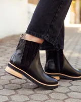 Botinetas tipo Chelsea de cuero negro, con paneles laterales elásticos y detalle de textura grabada en el talón. Presentan una suela plana con plataforma de madera clara y base de goma negra.