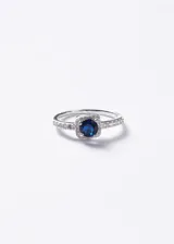 Anillo de plata 925 con piedra central de zirconia azul de corte redondo, enmarcada en una base cuadrada con pequeñas zirconias incrustadas en el aro.